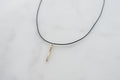 A thin black cord necklace with a small vertical gold-toned bar pendant on a white marble surface.