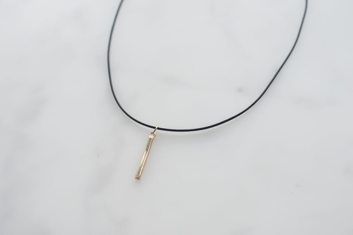 A thin black cord necklace with a small vertical gold-toned bar pendant on a white marble surface.