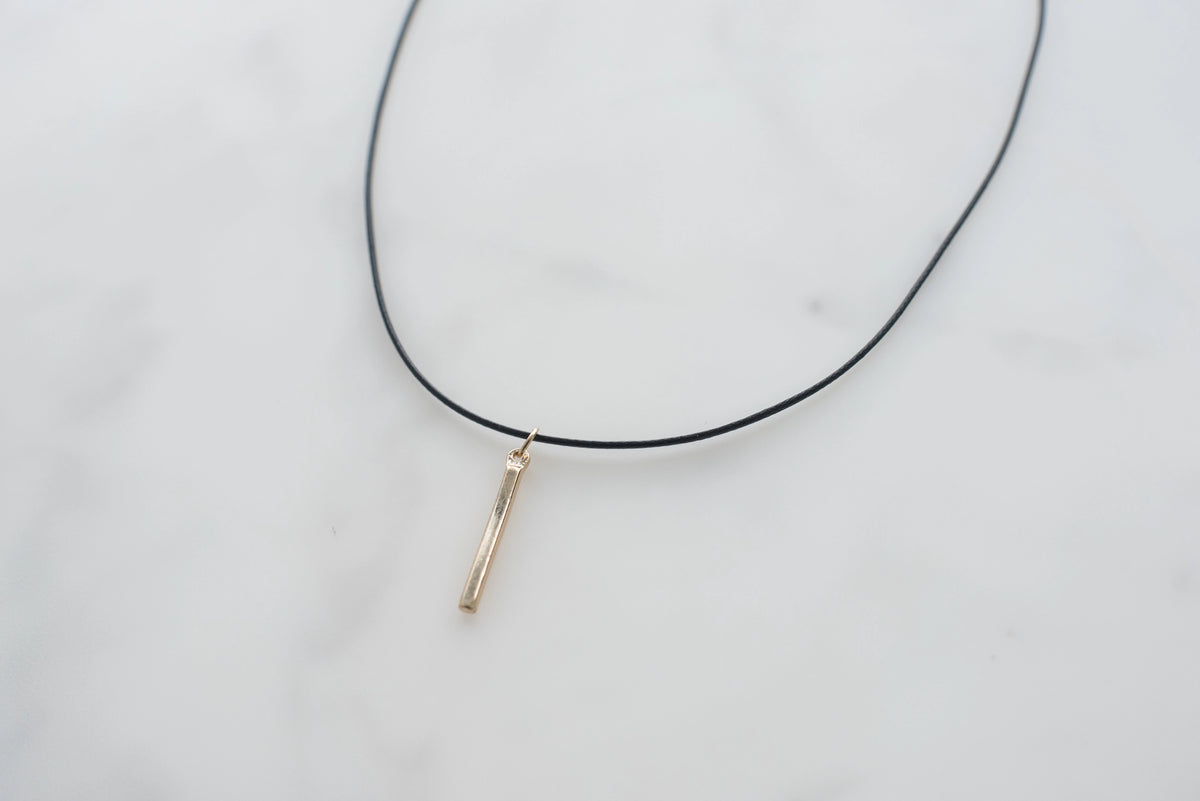 A thin black cord necklace with a small vertical gold-toned bar pendant on a white marble surface.