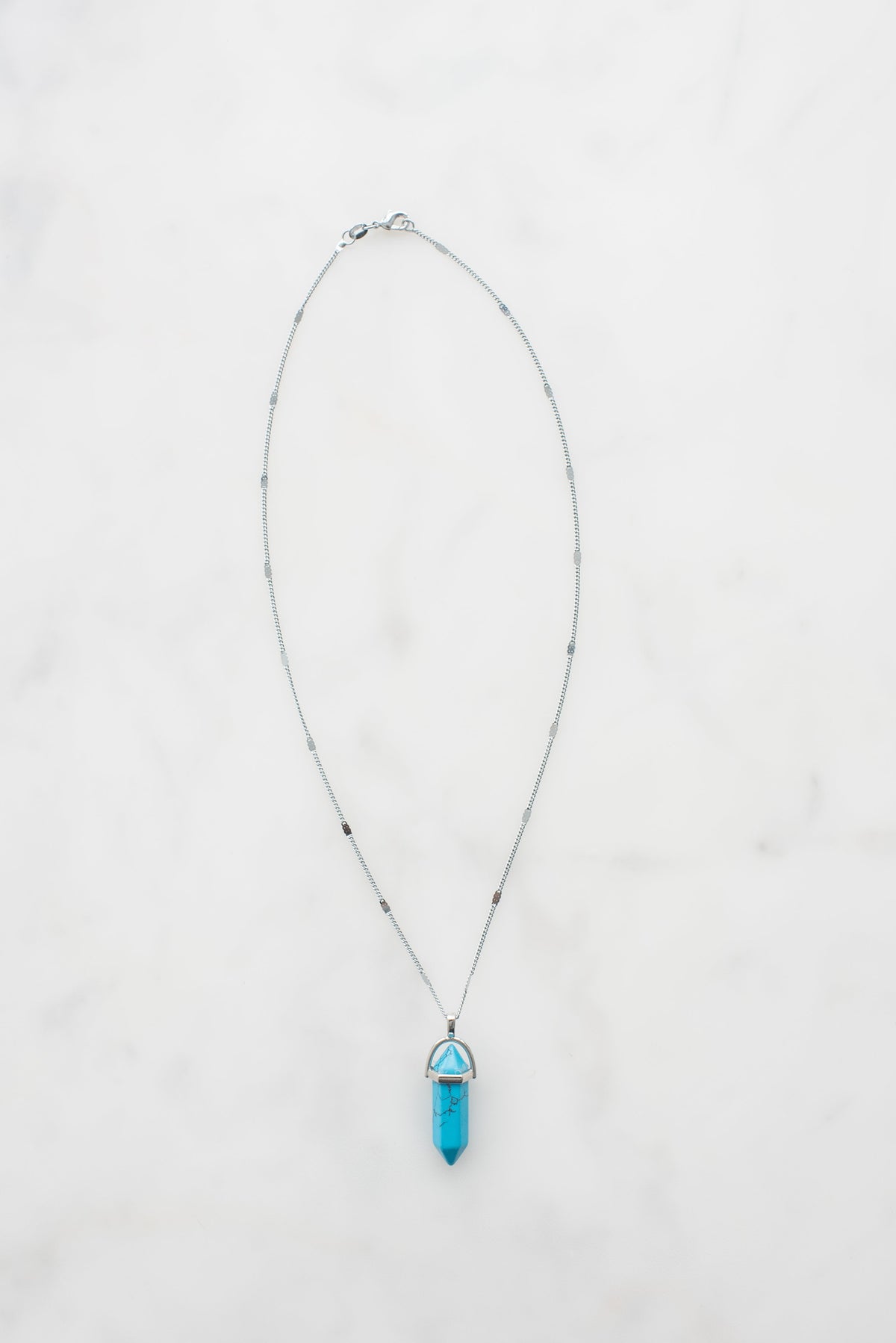 A silver chain necklace with a blue hexagonal crystal pendant on a white marble background.